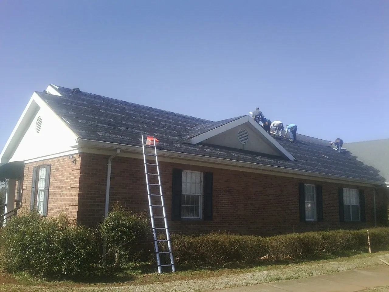 Crew tearing shingles off a brick ranch roof with a ladder set — Emergency Roof Repair work by our crew in Gattman
