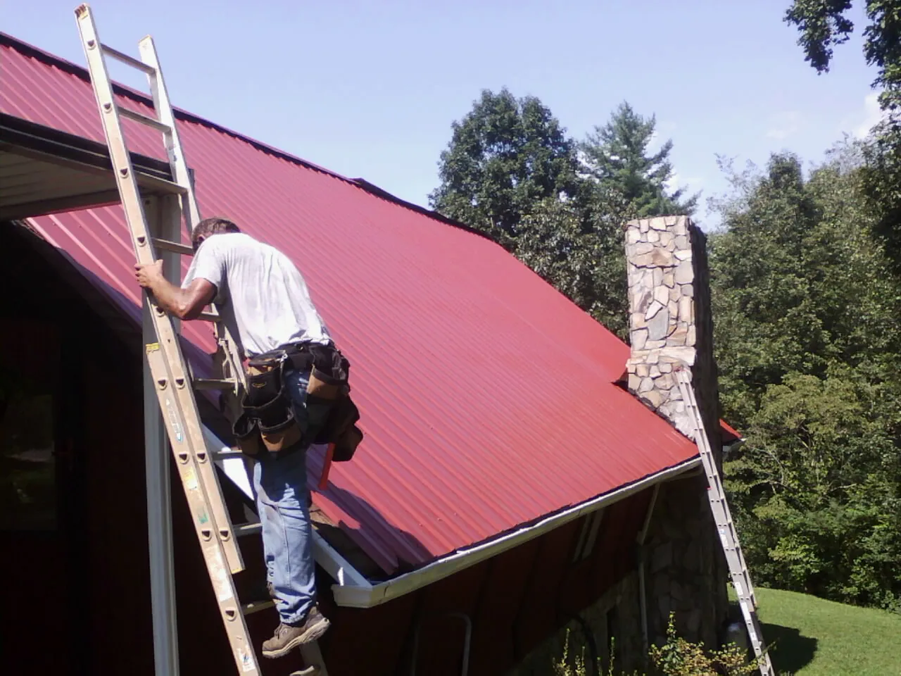 Roofer on a ladder beside a red metal roof and stone chimney — Soffit & Fascia Repair by Roofing Brothers in Gattman